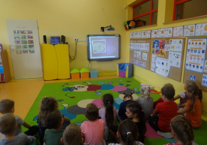 Dzieci siedzą na dywanie zwrócone w stronę tablicy interaktywnej i oglądają prezentację multimedialną na temat historii zegarów.